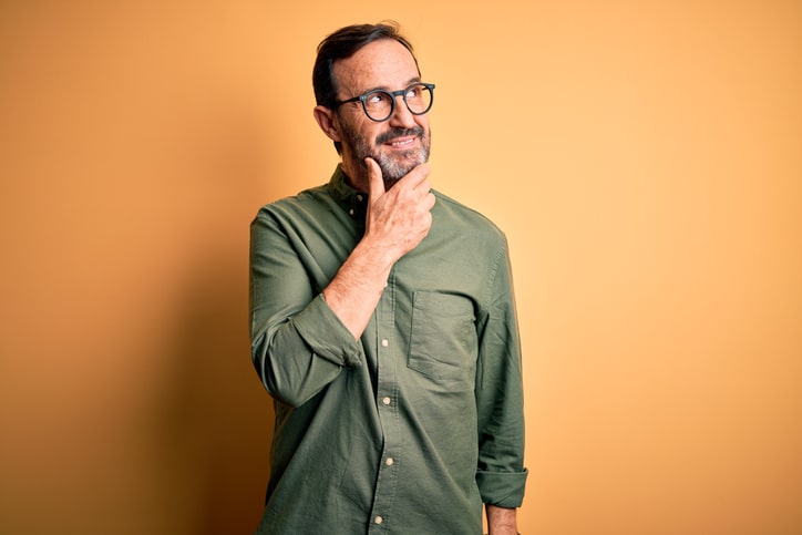 A middle aged man in gray glasses and a green button-up shirt stands against a orange background and holds his hand to his chin wondering about something with a smile on his face