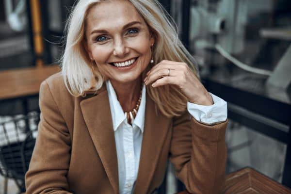 A mature woman with long blond hair smiles at the camera wearing a brown wool blazer and button up blouse