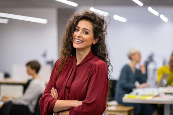 A young woman with curly hair in a professional red blouse smiles at the camera in an office setting