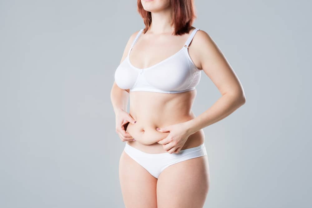 Postpartum woman standing in her underwear and pinching her stomach, in need of a mommy makover.